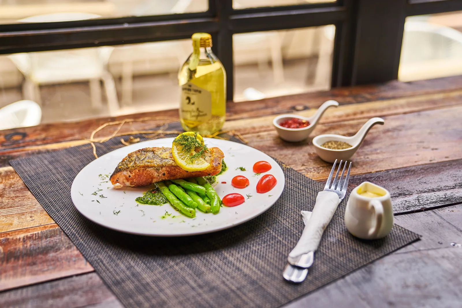 Cooked fish with asparagus, cherry tomatoes and lemon on a white plate beside a window.