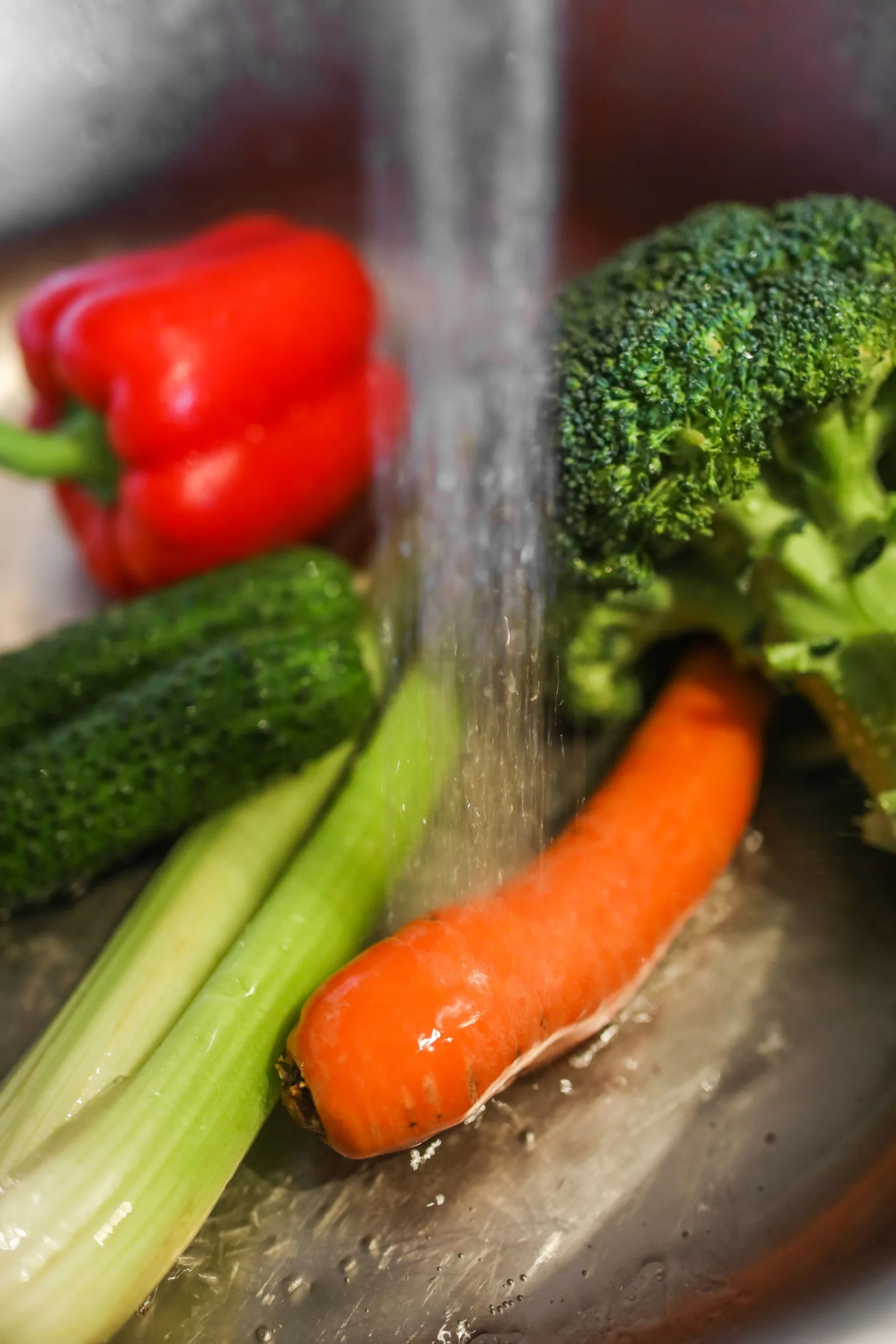 A stream of running water rinses carrots, broccoli, cucumber, celery and red pepper in a metal bowl.