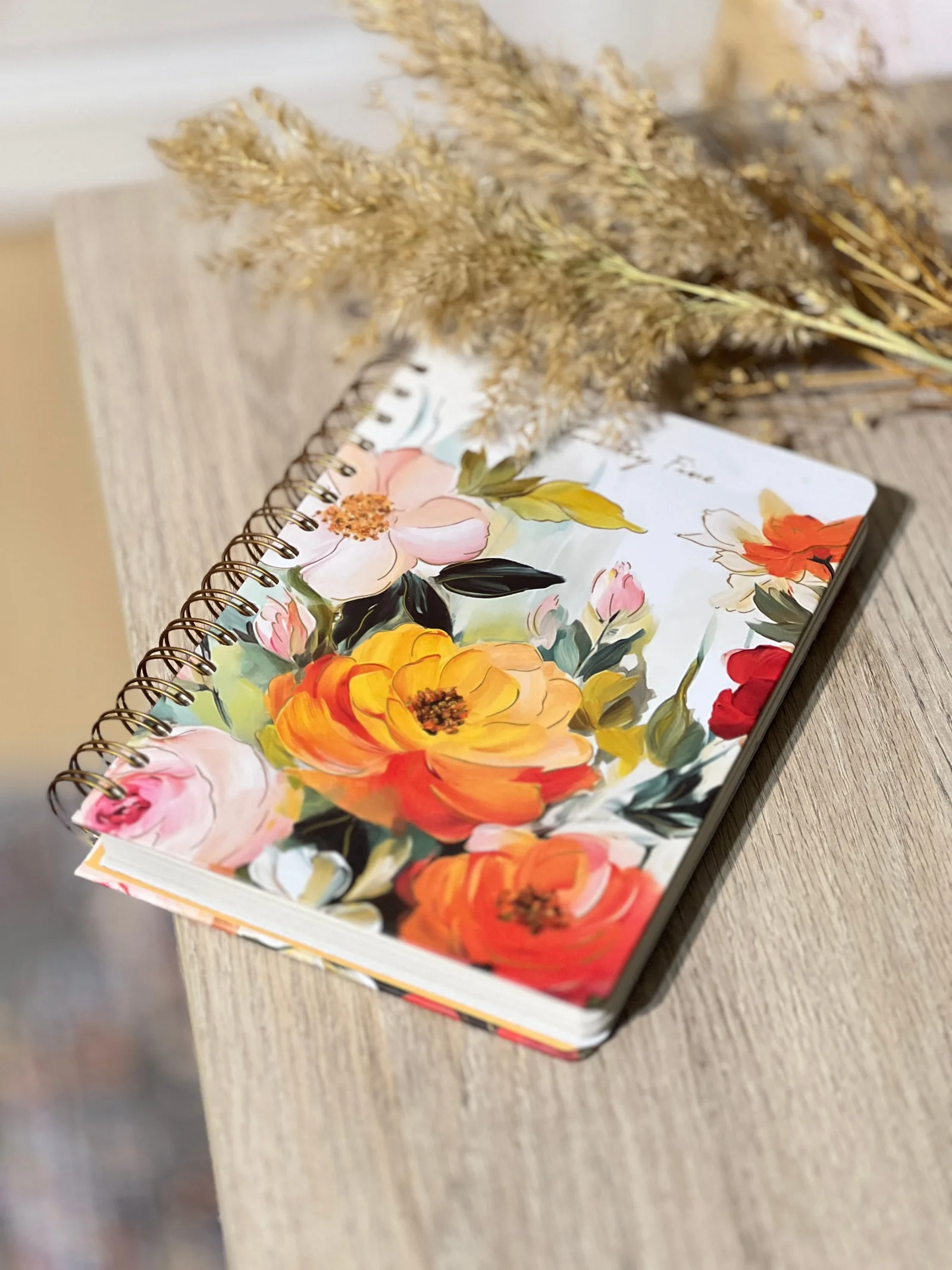A floral spiral notebook and dried stems on a wooden tabletop.