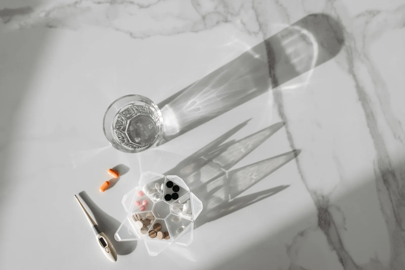 A glass of water, pill organizer and digital thermometer cast long morning shadows on a marble surface.