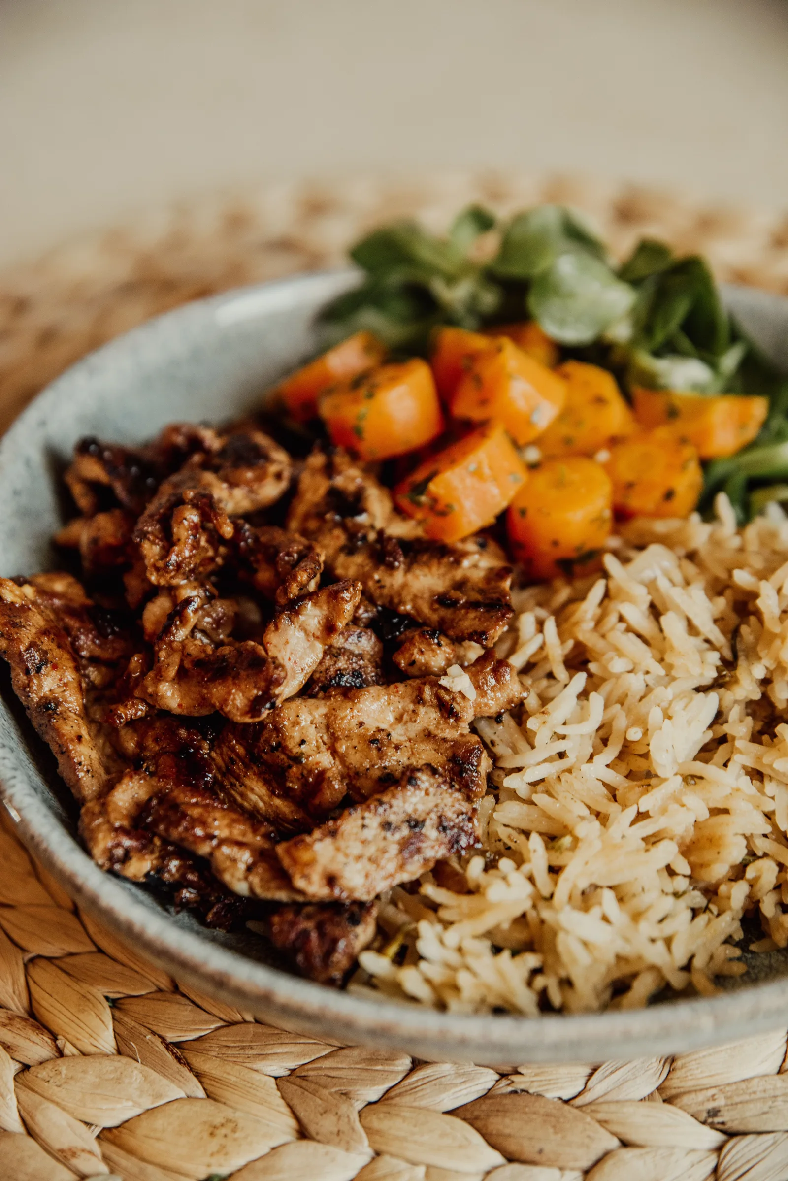 A bowl of grilled chicken, rice and colorful vegetables on a woven placemat.